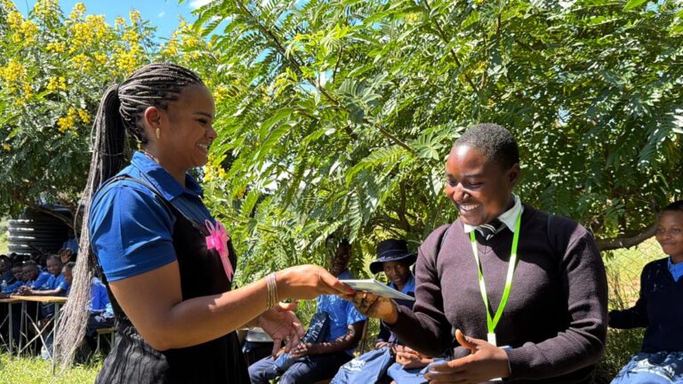 Leya Mnangagwa hands a book to a smiling student at a community visit, an example of purposeful business leadership in action.